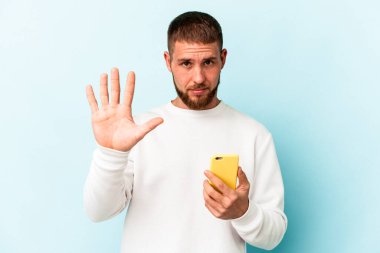 Elinde cep telefonu olan genç beyaz adam mavi arka planda izole edilmiş gülümseyen beş numarayı parmaklarıyla gösteriyor..