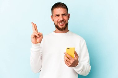 Elinde cep telefonu olan genç beyaz adam mavi arka planda izole edilmiş neşeli ve tasasız parmakları olan bir barış sembolü gösteriyor..