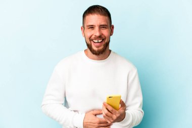 Mavi arka planda cep telefonu tutan genç beyaz adam gülüyor ve eğleniyor..