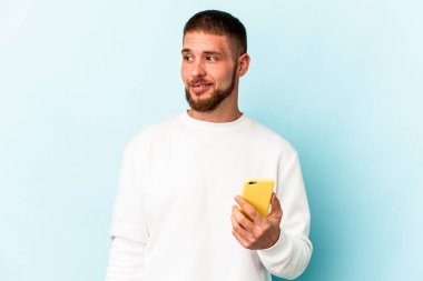 Mavi arka planda cep telefonu tutan genç beyaz adam gülümsüyor, neşeli ve keyifli görünüyor..