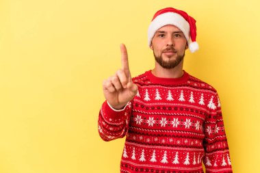 Genç beyaz adam, Noel 'i kutluyor. Sarı arka planda izole edilmiş. Parmağıyla bir numarayı gösteriyor..