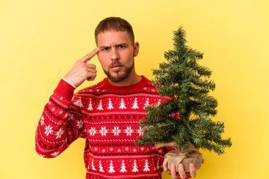 Genç beyaz adam Noel için küçük bir ağaç alıyor. Sarı arka planda izole edilmiş. İşaret parmağıyla yaptığı hayal kırıklığı jestini gösteriyor..