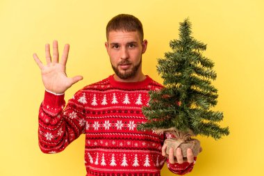 Sarı arka planda izole edilmiş küçük bir ağaç satın alan beyaz bir adam gülümseyerek beş numarayı parmaklarıyla gösteriyor..