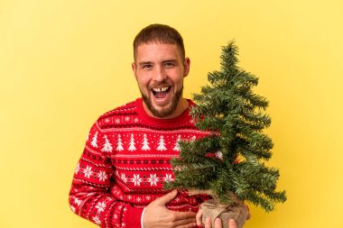 Genç beyaz adam Noel için küçük bir ağaç alıyor. Sarı arka planda izole edilmiş gülüyor ve eğleniyor..