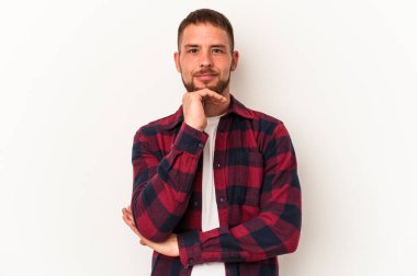 Beyaz arka planda izole edilmiş beyaz tenli genç beyaz adam mutlu ve kendinden emin gülümsüyor, elleriyle çenesine dokunuyor..