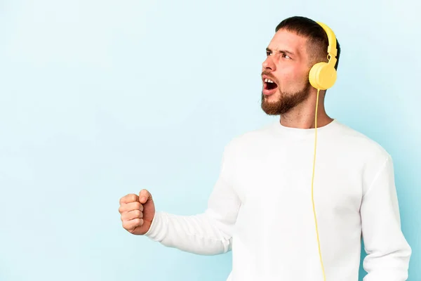 stock image Young caucasian man listening to music isolated on blue background raising fist after a victory, winner concept.