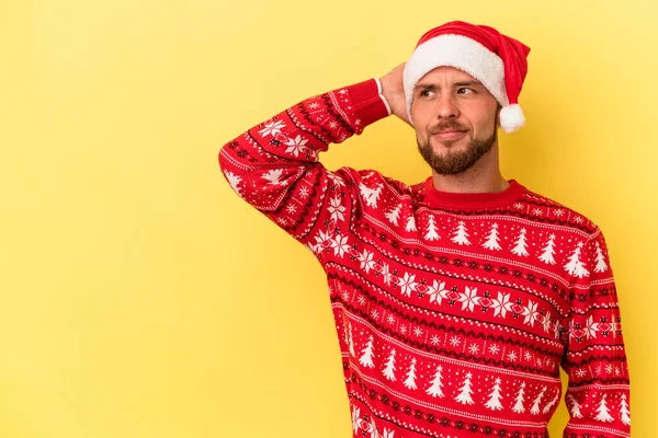 Kafkasyalı genç adam Noel 'i kutluyor sarı arka planda izole edilmiş kafasının arkasına dokunuyor, düşünüyor ve bir seçim yapıyor..