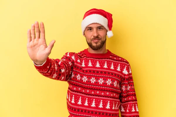 Genç, beyaz bir adam, sarı arka planda izole edilmiş bir şekilde Noel 'i kutluyor. Elini uzatmış, dur işareti gösteriyor..