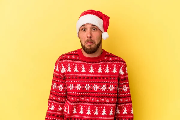 Kafkasyalı genç adam, sarı arka planda izole edilmiş Noel 'i kutluyor omuzlarını silkiyor ve gözleri açık..