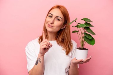 Pembe arka planda saklanmış bir bitki tutan beyaz bir kadın. Pembe arka planda izole edilmiş bir bitki tutan beyaz bir kadın. Parmağıyla bir numarayı gösteriyor..