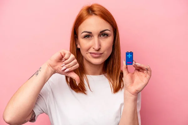 若い白人女性がピンクの背景に孤立した電池を持っていて 嫌なジェスチャー 親指を下に示しています 意見の相違 — ストック写真