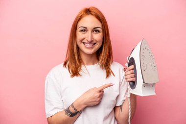 Pembe arka planda demir tutan genç beyaz kadın gülümsüyor ve boş alanda bir şeyler gösteriyor..
