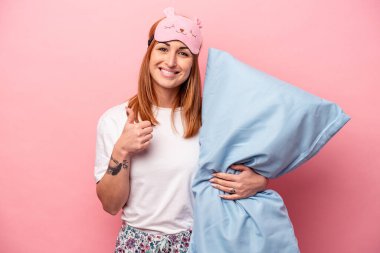 Pembe arka planda izole edilmiş pijama giyen beyaz bir kadın gülümsüyor ve başparmağını kaldırıyor.
