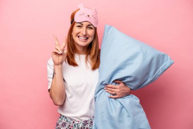 Pembe arka planda iki numarayı parmaklarıyla gösteren pijama giyen beyaz bir kadın..