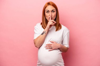 Genç, beyaz, hamile bir kadın pembe arka planda izole edilmiş. Sır saklıyor ya da sessizlik istiyor..