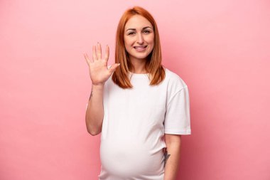 Genç, beyaz, hamile bir kadın pembe arka planda izole edilmiş. Gülümsüyor. Beş numarayı parmaklarıyla gösteriyor..