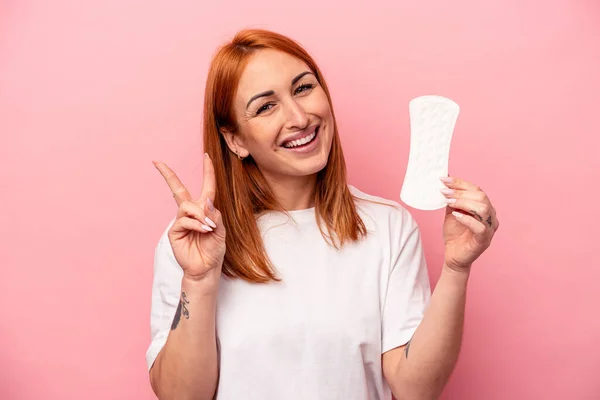ピンクの背景に孤立した生理用ナプキンを持つ若い白人女性喜びと屈託のない指で平和のシンボルを示す — ストック写真