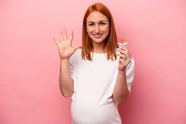 若いですCaucasian妊娠中の女性を保持ペースメーカー隔離上のピンクの背景笑顔陽気に示す番号5とともに指 — ストック写真