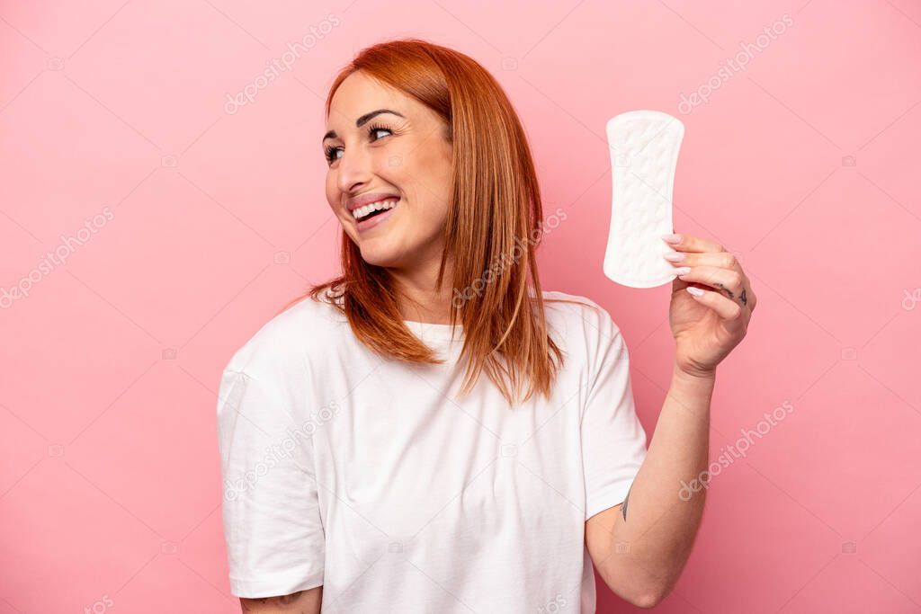 Young caucasian woman holding sanitary napkin isolated on pink background looks aside smiling, cheerful and pleasant.