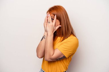 Beyaz arka planda izole edilmiş beyaz tenli genç bir kadın parmaklarının arasından korkmuş ve gergin bir şekilde göz kırpıyor..