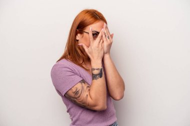 Beyaz arka planda izole edilmiş beyaz tenli genç bir kadın parmaklarının arasından korkmuş ve gergin bir şekilde göz kırpıyor..