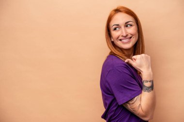 Beyaz tenli genç bir kadın bej arka planda izole edilmiş, başparmak uzakta, gülüyor ve kaygısız..