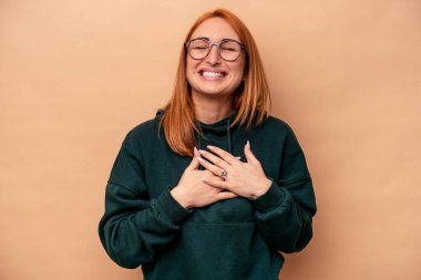 Beyaz tenli genç bir kadın bej arka planda izole edilmiş. Gülüyor, elini kalbe koyuyor, mutluluk kavramını koruyor..