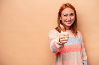 Beyaz tenli genç kadın bej arka planda izole edilmiş gülümsüyor ve başparmağını kaldırıyor.