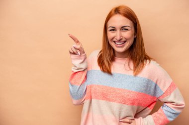 Beyaz tenli genç bir kadın, bej arka planda izole edilmiş. Gülümsüyor, işaret parmağıyla işaret ediyor..