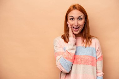 Beyaz tenli genç bir kadın bej arka planda izole edilmiş yüksek sesle bağırıyor, gözleri açık ve elleri gergin tutuyor..