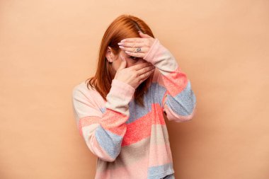 Beyaz tenli genç bir kadın bej arka planda izole edilmiş. Kameraya parmaklarıyla göz kırpıyor. Yüzünü kapatmaktan utanıyor..