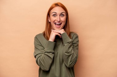 Beyaz tenli genç bir kadın bej arka planda izole edilmiş şans için dua ediyor, şaşırmış ve ağzını açıp önüne bakıyor..