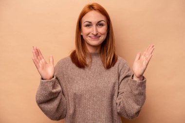 Beyaz tenli genç bir kadın, bej arka planda izole edilmiş. Elinde parmakları olan, gülümseyen ve kendinden emin bir şey var..