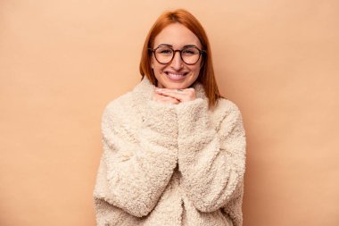 Beyaz tenli genç bir kadın bej arka planda izole edilmiş ellerini çenesinin altında tutuyor, mutlu bir şekilde yana bakıyor..