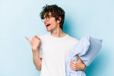 Genç beyaz bir adam pijama giyiyor ve yastık tutuyor. Mavi arka planda izole edilmiş. Baş parmak uzakta, güler yüzlü ve tasasız..