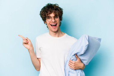 Genç beyaz adam pijama giyiyor ve yastığı mavi arka planda izole halde tutuyor gülümsüyor ve boş alanda bir şeyler gösteriyor..