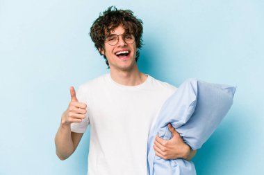 Genç beyaz bir adam pijama giyiyor ve yastık tutuyor mavi arka planda izole edilmiş gülümsüyor ve başparmağını kaldırıyor.