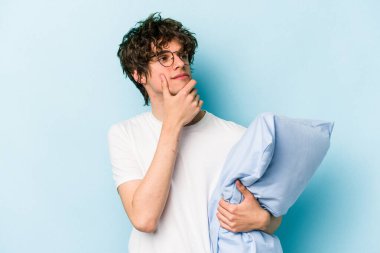 Pijama giyen ve yastık tutan genç beyaz adam mavi arka planda izole edilmiş şüpheli ve şüpheci bir ifadeyle yana bakıyor..