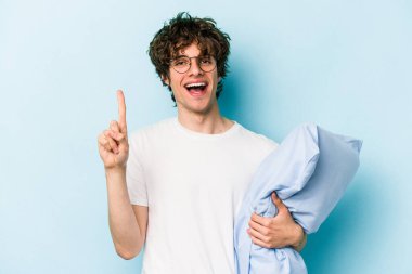 Genç, beyaz bir adam pijama giyiyor ve yastığı mavi arka planda tutuyor. Parmağıyla bir numarayı gösteriyor..