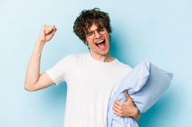 Pijama giyen ve yastık tutan genç beyaz adam mavi arka planda izole edilmiş zafer kazandıktan sonra yumruğunu kaldırıyor..