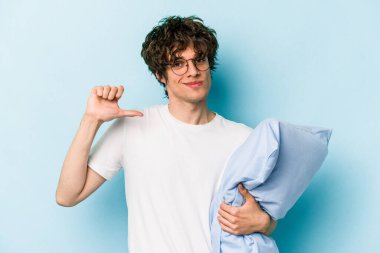 Genç beyaz adam pijama giyiyor ve yastığı mavi arka planda yalnız tutuyor gururlu ve kendinden emin, örnek olarak.