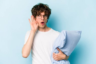 Genç beyaz bir adam pijama giyiyor ve yastık tutuyor. Mavi arka planda izole edilmiş bir şekilde bir dedikoduyu dinlemeye çalışıyor..