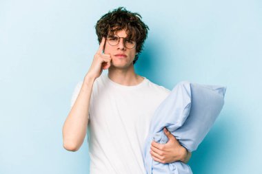 Genç, beyaz bir adam pijama giyiyor ve yastığı mavi arka planda izole edilmiş bir şekilde tutuyor. Şakağı parmağıyla işaret ediyor, düşünüyor ve bir göreve odaklanıyor..
