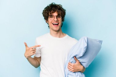 Pijama giyen ve mavi arka planda yastık tutan genç beyaz adam gömlek fotokopi alanını elle işaret ediyor, gururlu ve kendinden emin