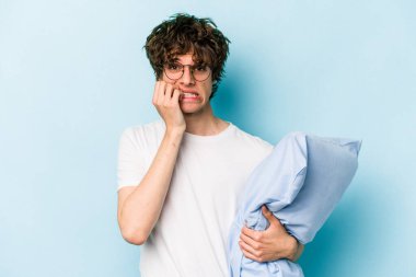 Genç, beyaz bir adam pijama giyiyor ve yastık tutuyor. Mavi arka planda izole edilmiş tırnak yiyor. Gergin ve endişeli..