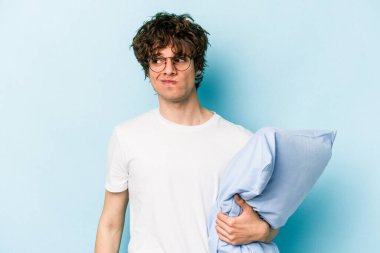 Genç beyaz bir adam pijama giyiyor ve yastığı mavi arka planda yalnız tutuyor. Kafası karışmış ve kendinden emin değil..