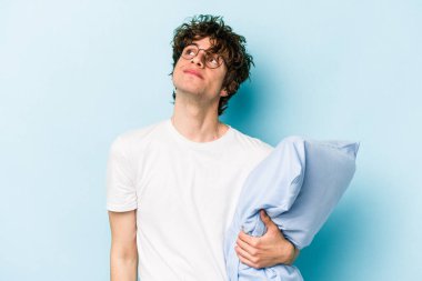 Genç, beyaz bir adam pijama giyiyor ve yastığı mavi arka planda tek başına tutuyor. Amaçlarına ve amaçlarına ulaşmayı hayal ediyor.