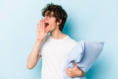 Genç beyaz bir adam pijama giyiyor ve yastığı mavi arka planda izole halde tutuyor bağırıyor ve avuç içini açık tutuyor..