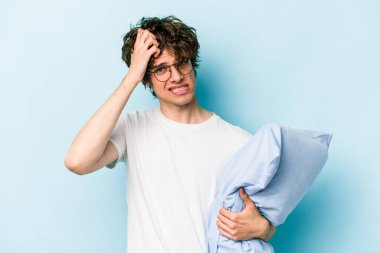 Mavi arka planda, pijama giyen ve yastık tutan genç beyaz adam şoka uğruyor..