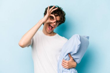 Genç beyaz bir adam pijama giyiyor ve yastığı mavi arka planda yalnız tutuyor..